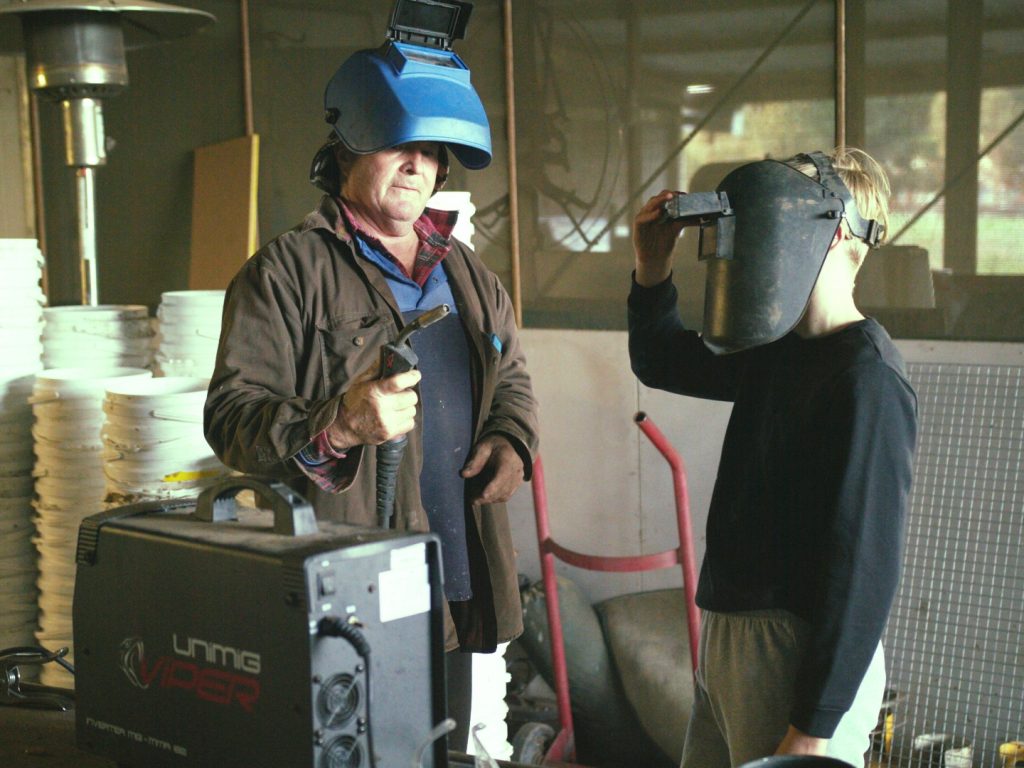 Man and boy wearing helmets with welding equipment in a workshop.