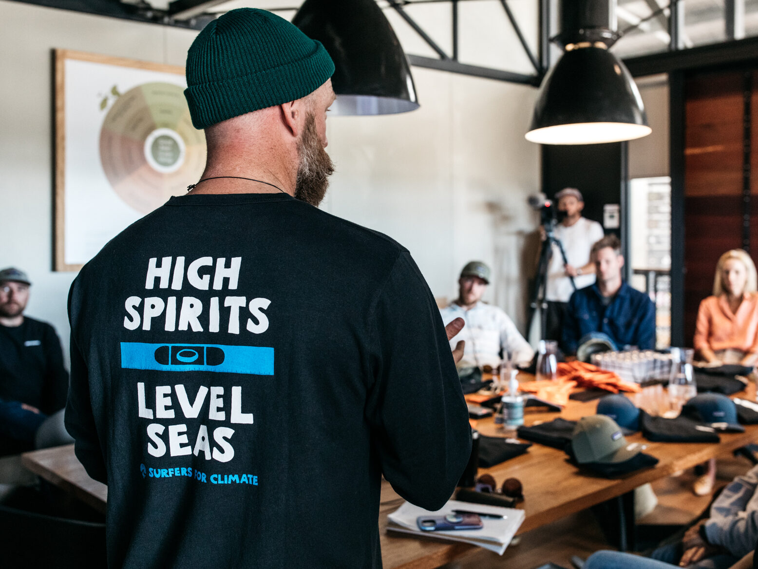 Man standing in front of a desk talking to people who are sitting down. His shirt says "High Spirits, Level Seas - Surfers for Climate"