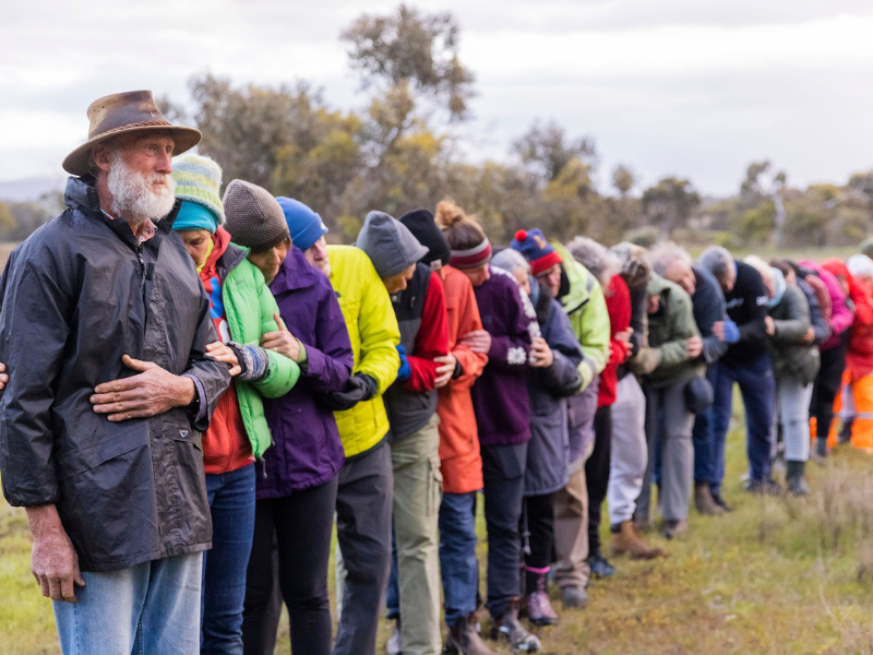 A group of people are in a line hugging each other like dominoes, wearing colourful clothing. 