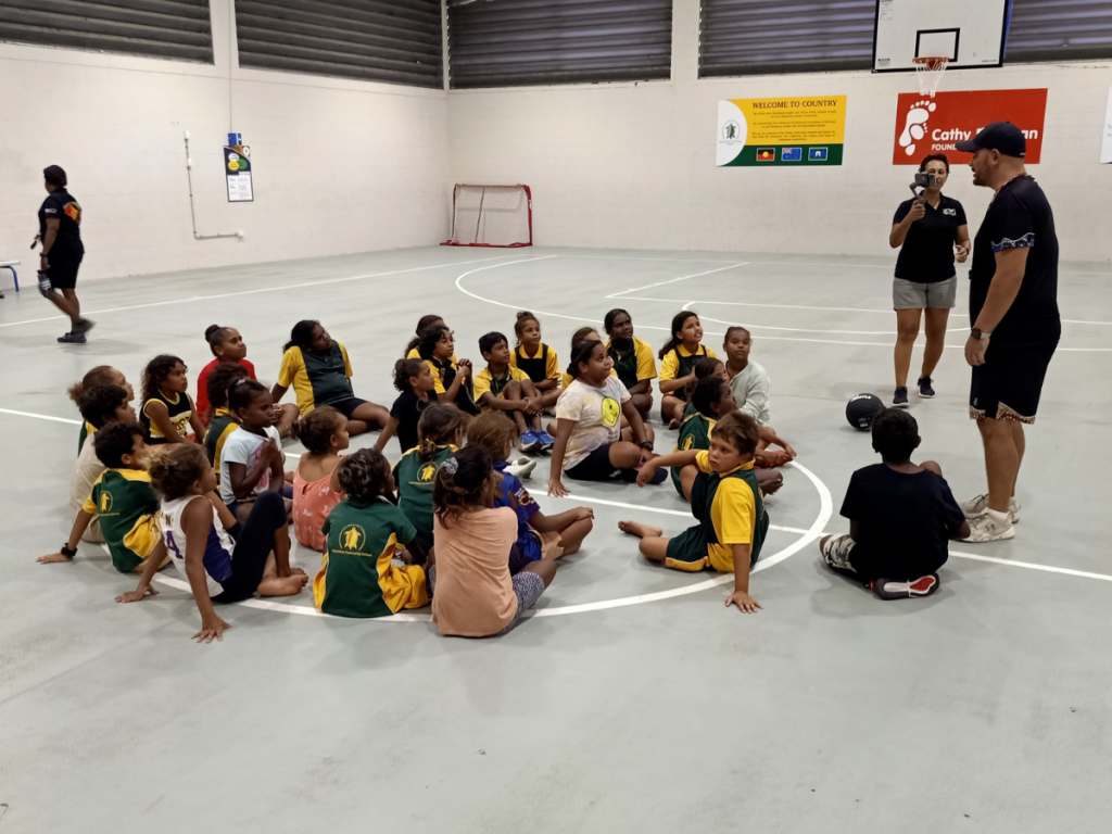 HEADING: 3x3 Basketball Brings Community Together. IMAGE: Large group of children listening to speaker