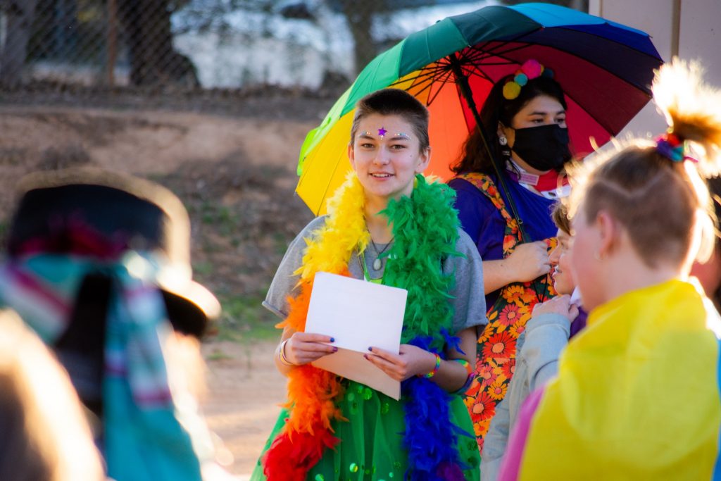 Riverland Youth Theatre member wearing a rainbow feather boa. There's someone else standing behind them with a rainbow umbrella.