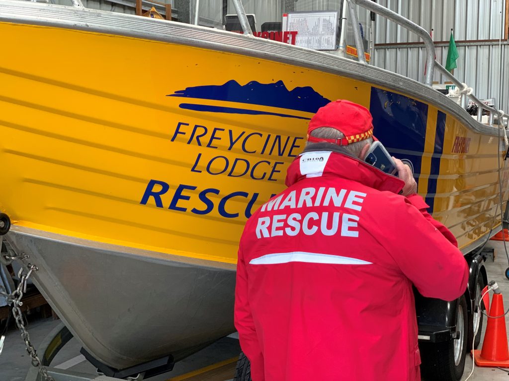 A man holding a mobile phone and wearing a red jacket that says Marine Rescue, standing in front of a yellow boat.
