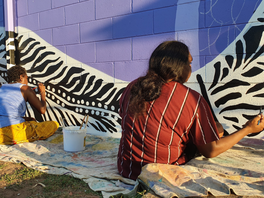 Artists sitting with their backs to the camera painting a mural on a wall.