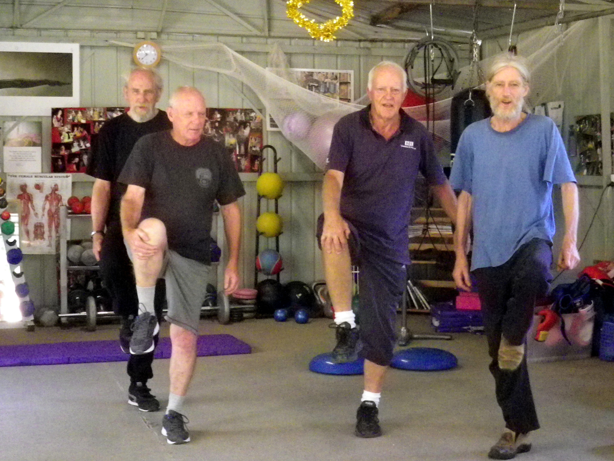 An exercise class, with four men standing one one leg.